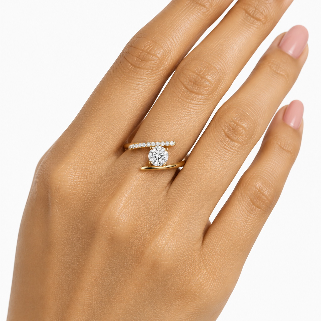 Female model's hand with warm golden Indian skin and soft warm nude-pink manicure wearing a very small yellow gold round brilliant diamond bypass solitaire ring — an open bypass design with two smooth plain polished yellow gold bar shanks crossing around a large round brilliant diamond solitaire in a four-prong yellow gold claw setting the upper bar set with round brilliant diamonds in shared pavé prong settings running along its length and the lower bar completely plain smooth polished yellow gold the two 