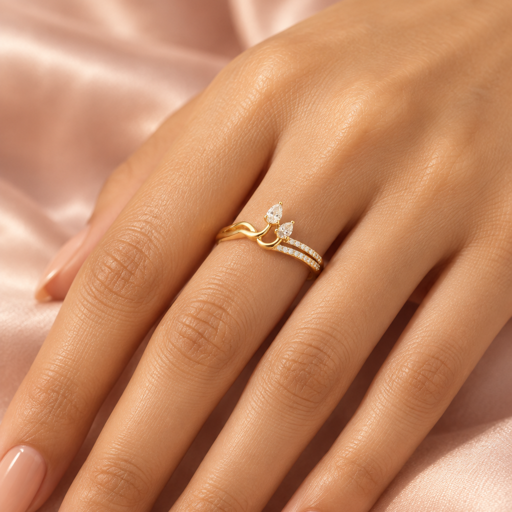 Female model's hand with warm light golden Indian skin and clean soft nude-peach manicure wearing a very small yellow gold sculptural asymmetric two-pear diamond ring — a fluid organic yellow gold band splitting and rising into an asymmetric sculptural knot setting at center with the plain smooth polished yellow gold shank curving upward in a fluid wave on the left and two pear-cut diamond solitaires in individual yellow gold claw prong settings emerging from the knot — one larger pear diamond angled upward