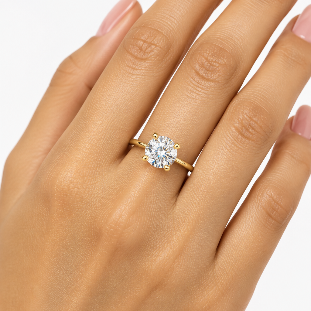 Female model's hand with warm light golden Indian skin and soft blush-pink manicure wearing a yellow gold round brilliant solitaire engagement ring featuring a slim plain high-mirror polished yellow gold band with a minimal low-profile four-prong basket setting with four small rounded ball-tipped yellow gold claw prongs holding a large round brilliant-cut diamond prominently above the band sparkling with exceptional brilliance fire and scintillation with flashes of white light and rainbow dispersion fingers