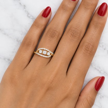 Female model's hand with warm medium Indian skin and classic red manicure wearing a very small yellow gold three-stone ring with a sweeping pavé diamond bypass frame featuring a slim plain yellow gold band sweeping into a wide horizontal elongated open lens-shaped frame — two curved pavé diamond rails fully set with continuous rows of small round brilliant diamonds in shared pavé prong settings forming a wide open eye shape with three round brilliant-cut diamonds in a horizontal row in individual four-prong