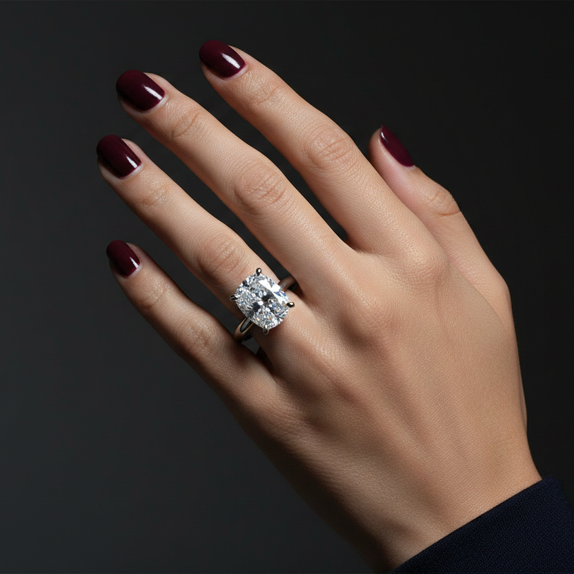Generated dramatic cinematic studio editorial variation - beautiful Indian female model wearing the white gold elongated cushion-cut diamond solitaire engagement ring, strong directional side light against near-black background with face-on overhead angle showing the full spectacular elongated cushion-cut diamond blazing with extraordinary icy crushed-ice brilliance and cool silver-white rim glow across the four ball-tip claws, deep red nails and dark navy sleeve