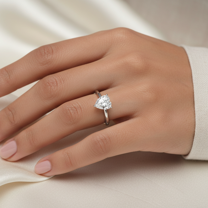 Generated image of a beautiful Indian female model wearing the white gold pear-cut diamond solitaire engagement ring with very large pear-cut diamond in elegant three-claw martini setting with one ball-tip claw at the pointed tip and two ball-tip claws cradling the rounded base on a smooth slim plain white gold shank — the ultimate pure and elegant pear solitaire