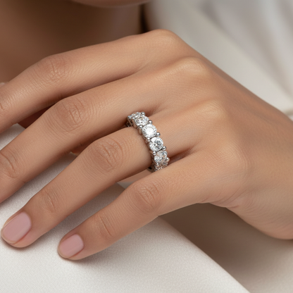 Generated image of a beautiful Indian female model wearing the white gold seven-stone round brilliant diamond half eternity band — seven large round brilliant diamonds blazing simultaneously with extraordinary icy white fire and rainbow scintillation in their elegant shared-prong four-claw white gold settings running in a graceful arc across the top of the smooth wide plain white gold shank, soft cool diffused studio lighting on neutral background with elegant three-quarter angle pose
