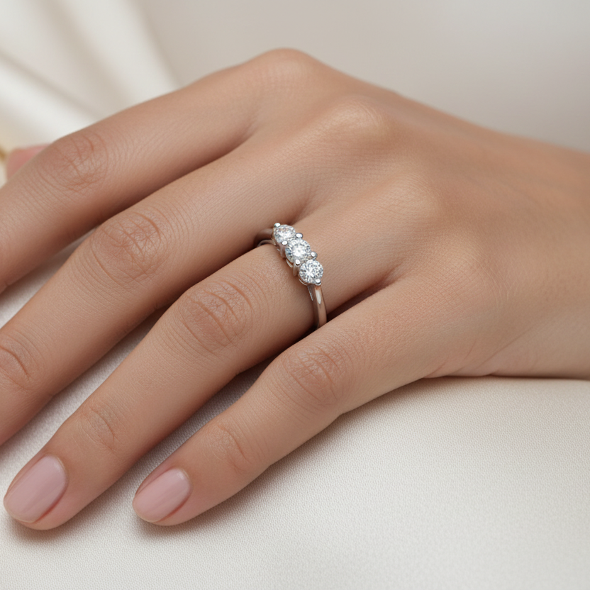 Generated image of a beautiful Indian female model wearing the white gold three-stone round brilliant diamond trilogy ring with three equal-sized round brilliant diamonds in beautiful shared flat-bar white gold claw settings sitting closely together in a tight three-diamond cluster on a smooth wide plain white gold shank all blazing simultaneously with extraordinary icy white fire and rainbow scintillation