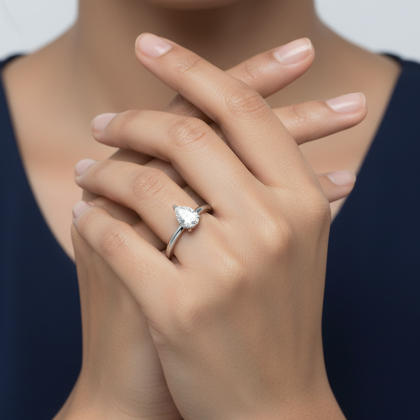 Generated image of an Indian female model wearing the white gold pear-cut diamond solitaire engagement ring with three-claw open basket setting and slim plain polished shank