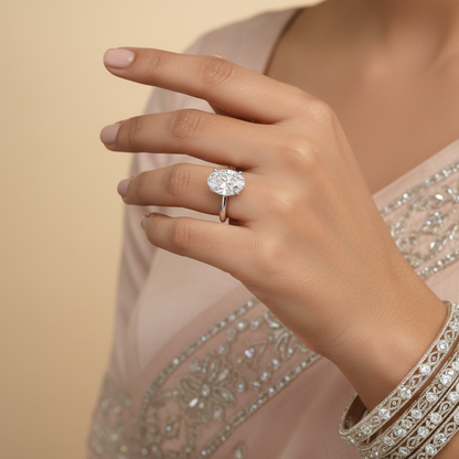 Generated Indian bridal editorial variation - beautiful Indian female model wearing the white gold oval-cut diamond solitaire engagement ring with spectacular very large oval-cut diamond oriented north-south along the finger, warm studio lighting on champagne background with soft ivory silk saree blouse with delicate silver embroidery, soft rose nails and delicate diamond or pearl bangles stacked alongside for a beautiful traditional Indian bridal context