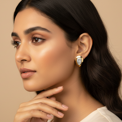 Generated very small dainty size variation - beautiful Indian female model wearing the yellow gold pear-cut diamond solitaire stud earrings with wide flat teardrop bezel frame as extremely tiny delicate petite accent studs, warm golden hour light on champagne background with soft romantic waves hairstyle and ivory silk outfit
