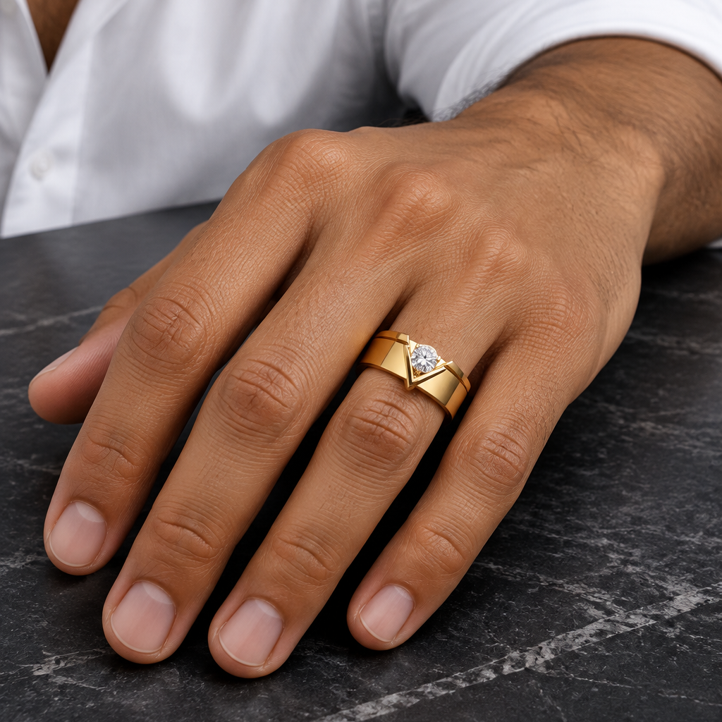 Handsome elegant Indian male model's hand with warm golden skin and well-groomed clean short nails wearing a very small wide yellow gold men's solitaire ring with a bold V-chevron design — wide flat polished yellow gold band with a prominent V-shaped chevron cutout or groove on the face pointing downward with a round brilliant diamond in a low bezel tension-style yellow gold setting nestled at the point of the V; hand resting confidently on a smooth dark grey slate or black marble surface fingers relaxed an
