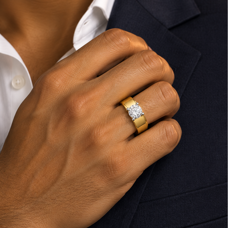Handsome elegant male model with warm medium golden Indian skin and well-groomed hands wearing a very small yellow gold round brilliant diamond solitaire wide band ring — a wide thick smooth plain polished yellow gold band with a broad flat architectural profile and a large round brilliant diamond solitaire in a four-prong white gold claw setting elevated on a low cathedral mount ring worn on the ring finger hand resting casually with a relaxed confident grip holding the lapel of a dark navy charcoal suit j