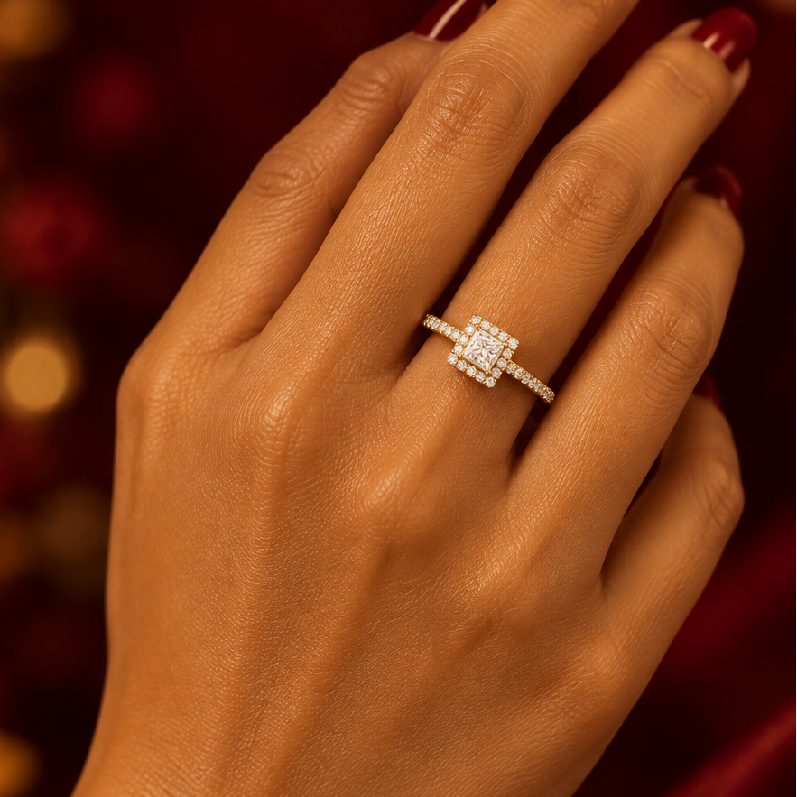 Indian female model's hand with warm deep golden skin and classic deep red crimson manicure wearing a very small yellow gold princess-cut diamond halo engagement ring — a slim yellow gold pavé diamond band with round brilliant diamonds along both sides and a square princess-cut diamond solitaire in a four-prong yellow gold setting surrounded by a square halo frame of round brilliant diamonds creating a glittering square halo silhouette; hand elegantly raised with the back of the hand toward the camera showi