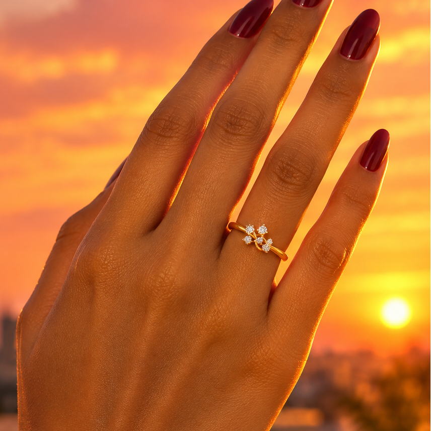 Indian female model's hand with warm golden skin and bold rich burgundy deep wine manicure wearing a very small yellow gold diamond crossover bypass ring — a slim plain polished yellow gold shank with five round brilliant diamonds in three-prong martini settings scattered asymmetrically in a loose casual constellation cluster at varying heights; hand raised against a glowing warm sunset sky fingers elegantly extended and slightly apart the ring catching warm orange-gold sunset light making the five scattere