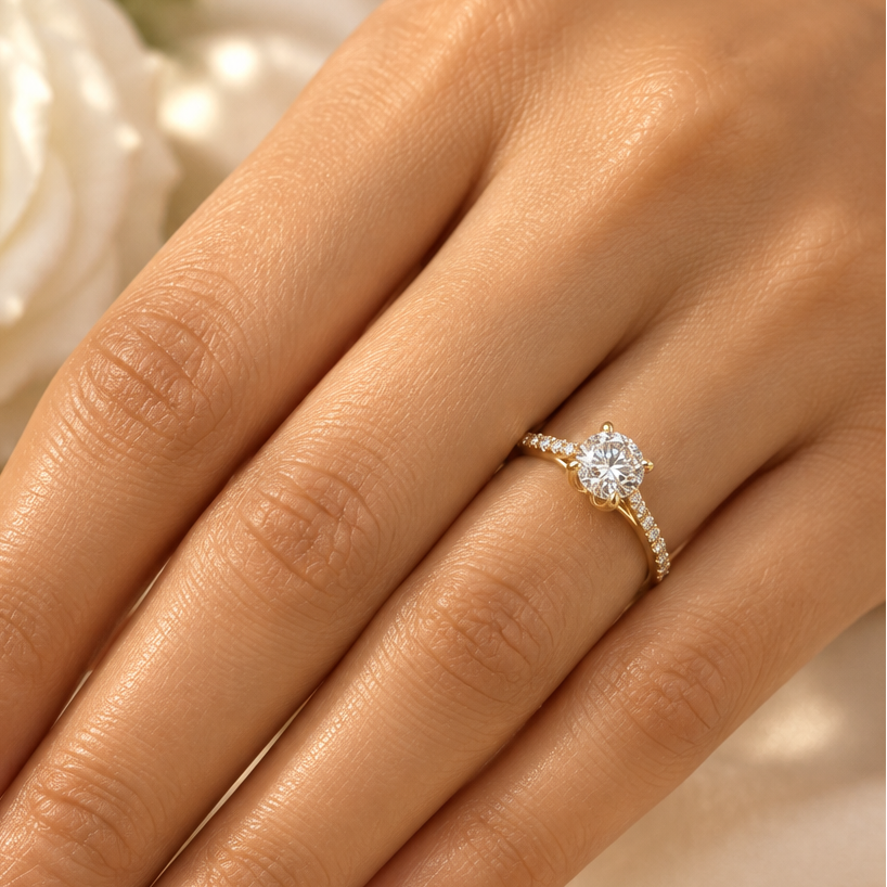 Indian female model's hand with warm golden skin and classic soft blush-nude pale rose manicure wearing a very small yellow gold round brilliant diamond solitaire engagement ring with pavé diamond shoulders — a slim yellow gold band with round brilliant diamonds in pavé settings running along both shoulders rising to a four-prong yellow gold tulip double-claw setting elevating a large round brilliant diamond solitaire prominently above the band blazing with maximum fire and scintillation; ring worn alone on