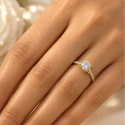 Indian female model's hand with warm golden skin and classic soft blush-nude pale rose manicure wearing a very small yellow gold round brilliant diamond solitaire engagement ring with pavé diamond shoulders — a slim yellow gold band with round brilliant diamonds in pavé settings running along both shoulders rising to a four-prong yellow gold tulip double-claw setting elevating a large round brilliant diamond solitaire prominently above the band blazing with maximum fire and scintillation; ring worn alone on