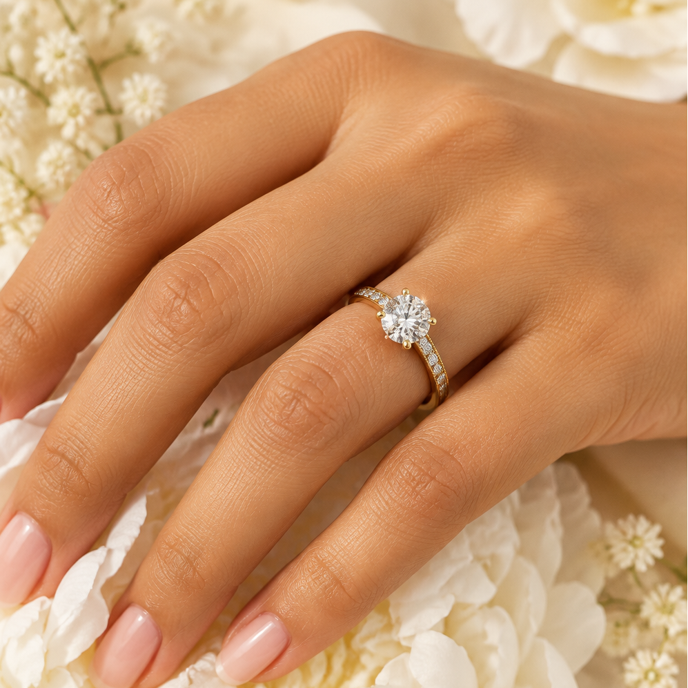 Indian female model's hand with warm golden skin and classic soft blush-pink manicure wearing a very small yellow gold round brilliant diamond solitaire engagement ring with a diamond channel band — a yellow gold shank with a row of round brilliant diamonds running along both shoulders in channel shared prong settings with delicate milgrain edge detail; the shoulders rising to a four-prong yellow gold claw setting elevating a large round brilliant diamond solitaire prominently above the band blazing with br