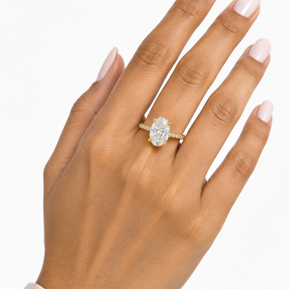 Indian female model's hand with warm golden skin and clean soft ivory pale blush-white manicure wearing a very small yellow gold oval-cut diamond solitaire engagement ring with a pavé diamond band — a slim yellow gold band with round brilliant diamonds in pavé settings running along both shoulders rising to a four-prong yellow gold claw setting elevating a large elongated oval-cut diamond solitaire prominently above the band set with its long axis along the finger; ring worn alone on the ring finger hand el