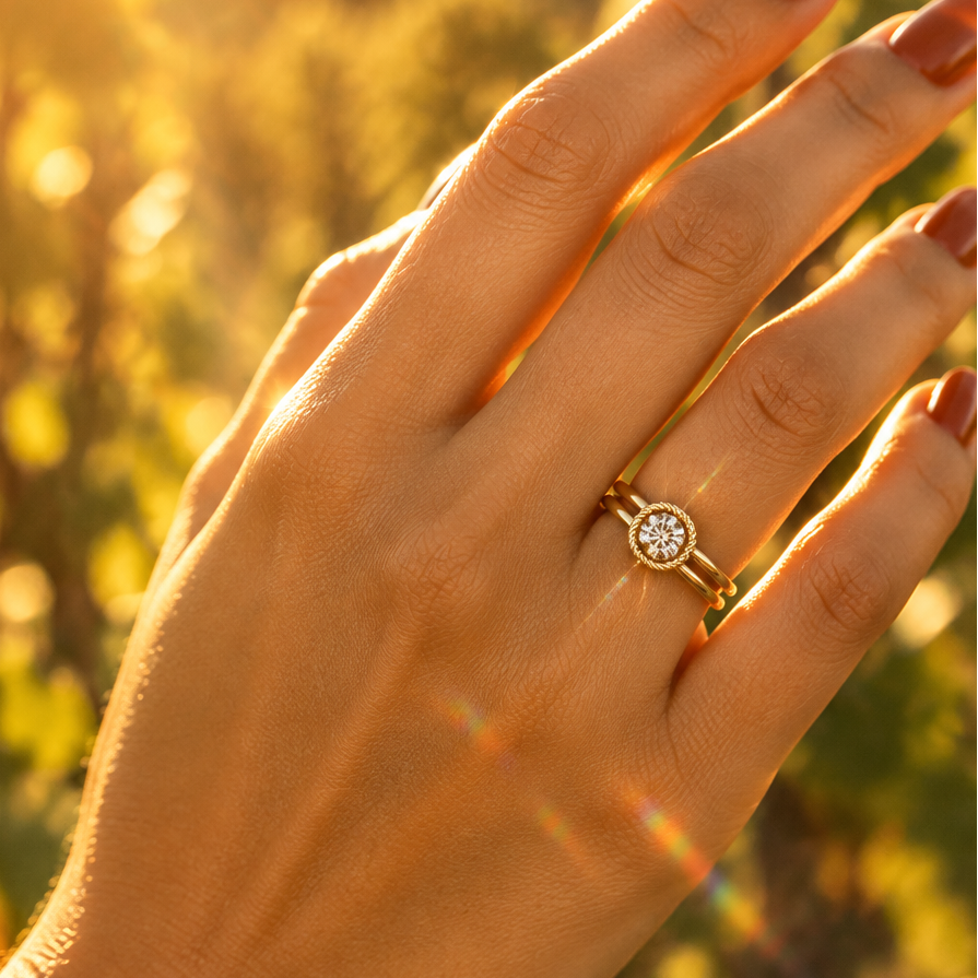 Indian female model's hand with warm golden skin and deep rich terracotta burnt orange manicure wearing a very small yellow gold round brilliant diamond solitaire double-shank rope bezel ring — a split double-band shank with two parallel smooth rounded plain polished yellow gold bands converging at the center top into a circular round bezel frame with a decorative twisted rope braided yellow gold border within which a large round brilliant diamond solitaire sparkles brilliantly catching warm golden hour lig
