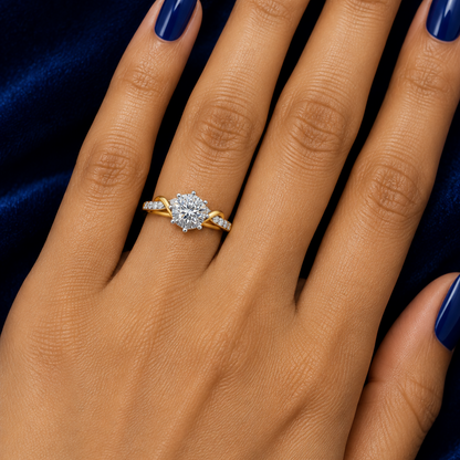 Indian female model's hand with warm golden skin and deep royal blue sapphire manicure wearing a very small yellow gold round brilliant diamond solitaire ring with a swirl bypass diamond band — dramatic yellow gold shank sweeping up on both sides in a flowing S-curve scroll wave design beneath the center stone with channel-set round brilliant diamonds along both the yellow gold and white gold portions; large round brilliant diamond in a six-prong silver white gold crown setting with rounded silver prong tip