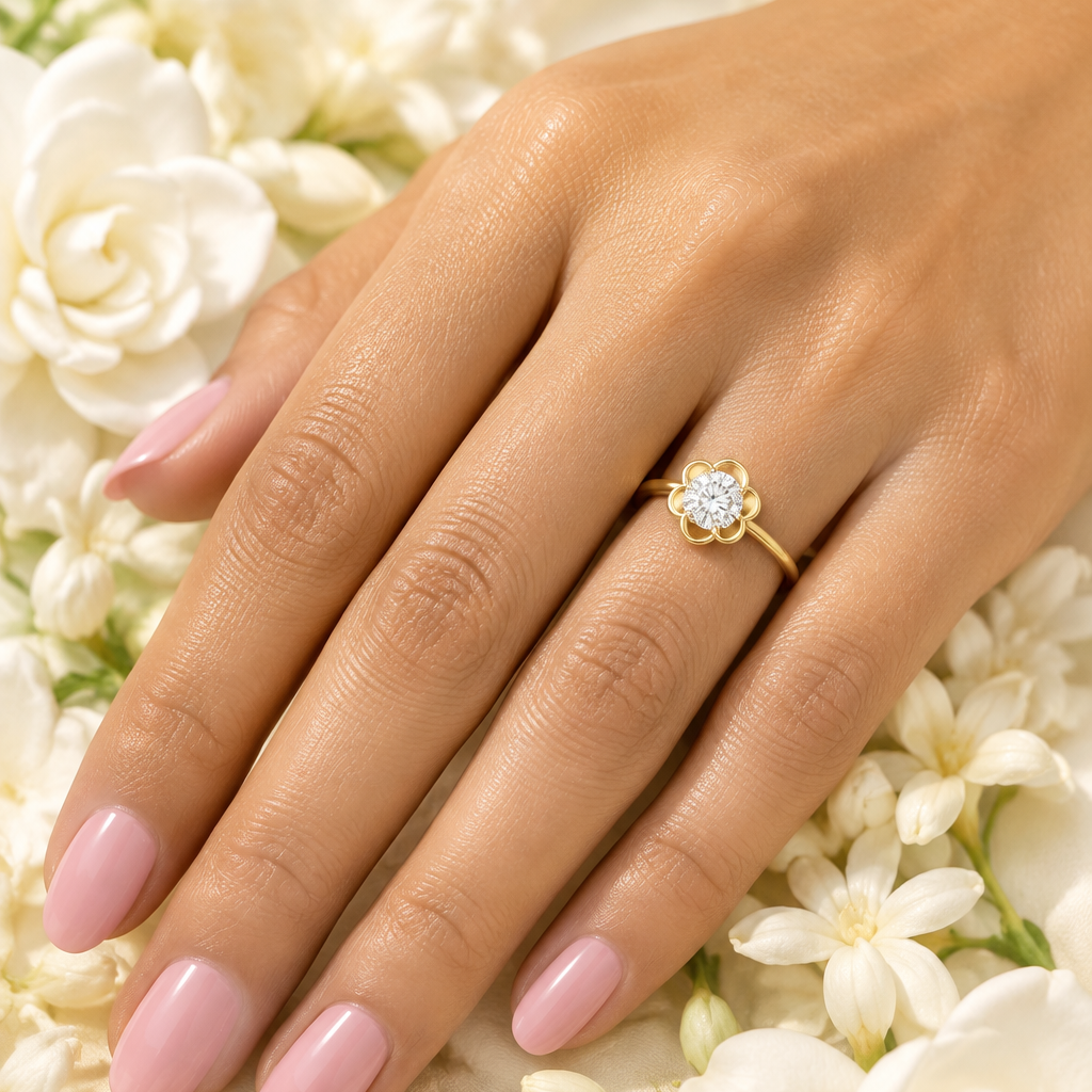 Indian female model's hand with warm golden skin and soft blush petal pink manicure wearing a very small yellow gold round brilliant diamond solitaire flower bezel ring — a slim plain polished yellow gold band rising to a large round brilliant diamond solitaire set within a scalloped four-petal flower-shaped yellow gold bezel frame with four gracefully curved rounded petal lobes radiating symmetrically outward surrounding the diamond like a blooming flower each petal lobe a smooth polished yellow gold curve