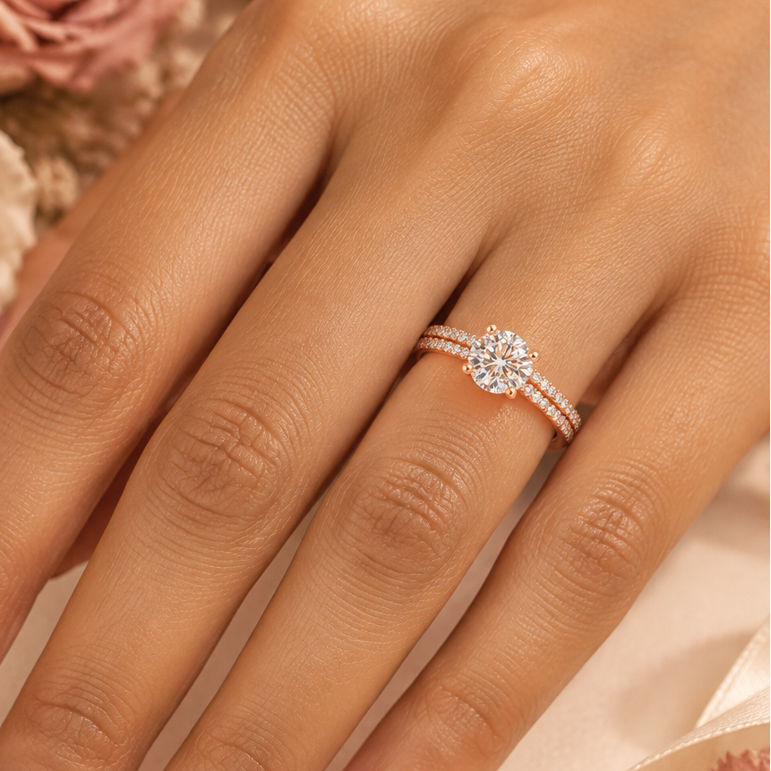 Indian female model's hand with warm golden skin and soft warm dusty rose antique rose manicure wearing a very small rose gold round brilliant diamond solitaire engagement ring with a double-row pavé diamond band — rose gold band with two parallel rows of round brilliant diamonds in pavé settings running along both shoulders creating a rich full-diamond double-row shoulder effect; the shoulders rising to a four-prong rose gold ball-tip claw setting holding a large round brilliant diamond solitaire blazing w