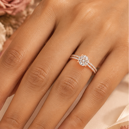 Indian female model's hand with warm golden skin and soft warm dusty rose antique rose manicure wearing a very small rose gold round brilliant diamond solitaire engagement ring with a double-row pavé diamond band — rose gold band with two parallel rows of round brilliant diamonds in pavé settings running along both shoulders creating a rich full-diamond double-row shoulder effect; the shoulders rising to a four-prong rose gold ball-tip claw setting holding a large round brilliant diamond solitaire blazing w