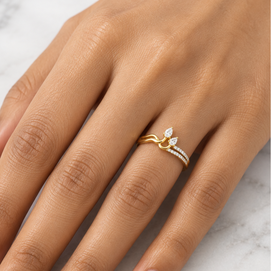 Indian female model's hand with warm golden skin and soft warm nude pale champagne-pink manicure wearing a very small yellow gold sculptural two-pear diamond ring — a slim yellow gold shank with a split bypass design: one side a plain polished yellow gold wave band the other side carrying a diagonal row of round brilliant diamonds in pavé running toward the top; at the top two pear-shaped diamonds nestled closely together like butterfly wings petal duo in a toi et moi inspired arrangement one pear slightly 