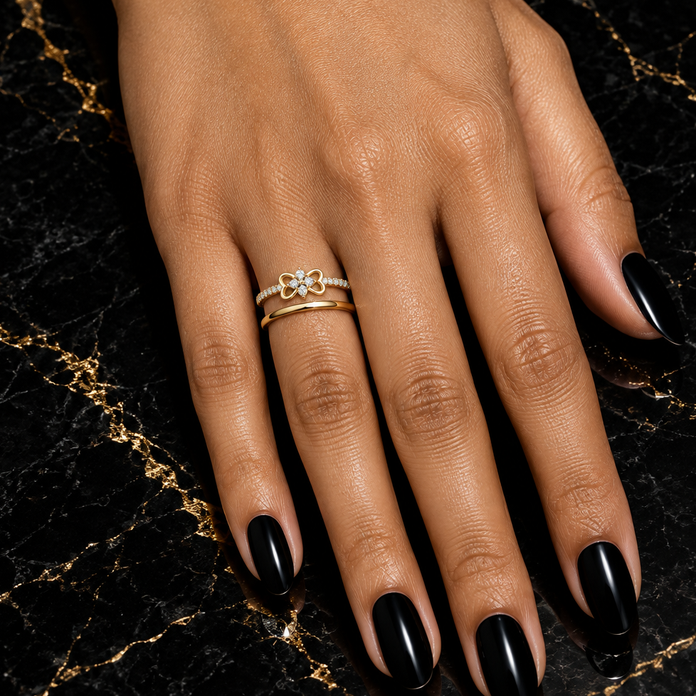 Indian female model's hand with warm golden skin and striking bold black manicure wearing a very small yellow gold four-leaf clover diamond ring — a slim yellow gold pavé shank with round brilliant diamonds along both sides and a delicate four-leaf clover motif in plain polished yellow gold with four small open heart-shaped petal loops radiating symmetrically outward and a small tight cluster of round brilliant diamonds at center; ring worn alongside one slim plain yellow gold stacking band; hand elegantly 