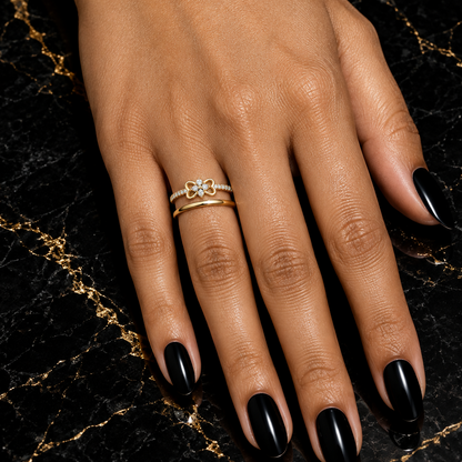 Indian female model's hand with warm golden skin and striking bold black manicure wearing a very small yellow gold four-leaf clover diamond ring — a slim yellow gold pavé shank with round brilliant diamonds along both sides and a delicate four-leaf clover motif in plain polished yellow gold with four small open heart-shaped petal loops radiating symmetrically outward and a small tight cluster of round brilliant diamonds at center; ring worn alongside one slim plain yellow gold stacking band; hand elegantly 