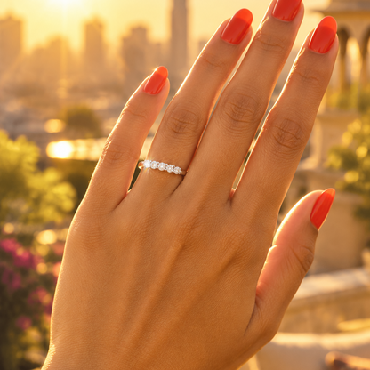 Indian female model's hand with warm golden skin and striking bold coral vibrant orange-red manicure wearing a very small white gold platinum round brilliant diamond half-eternity band — a smooth plain polished white gold platinum shank with seven round brilliant diamonds in shared U-shaped prong settings running in a continuous line across the top creating a seamless river of diamond brilliance; the eternity band worn alone on the ring finger hand raised confidently with fingers naturally spread and slight